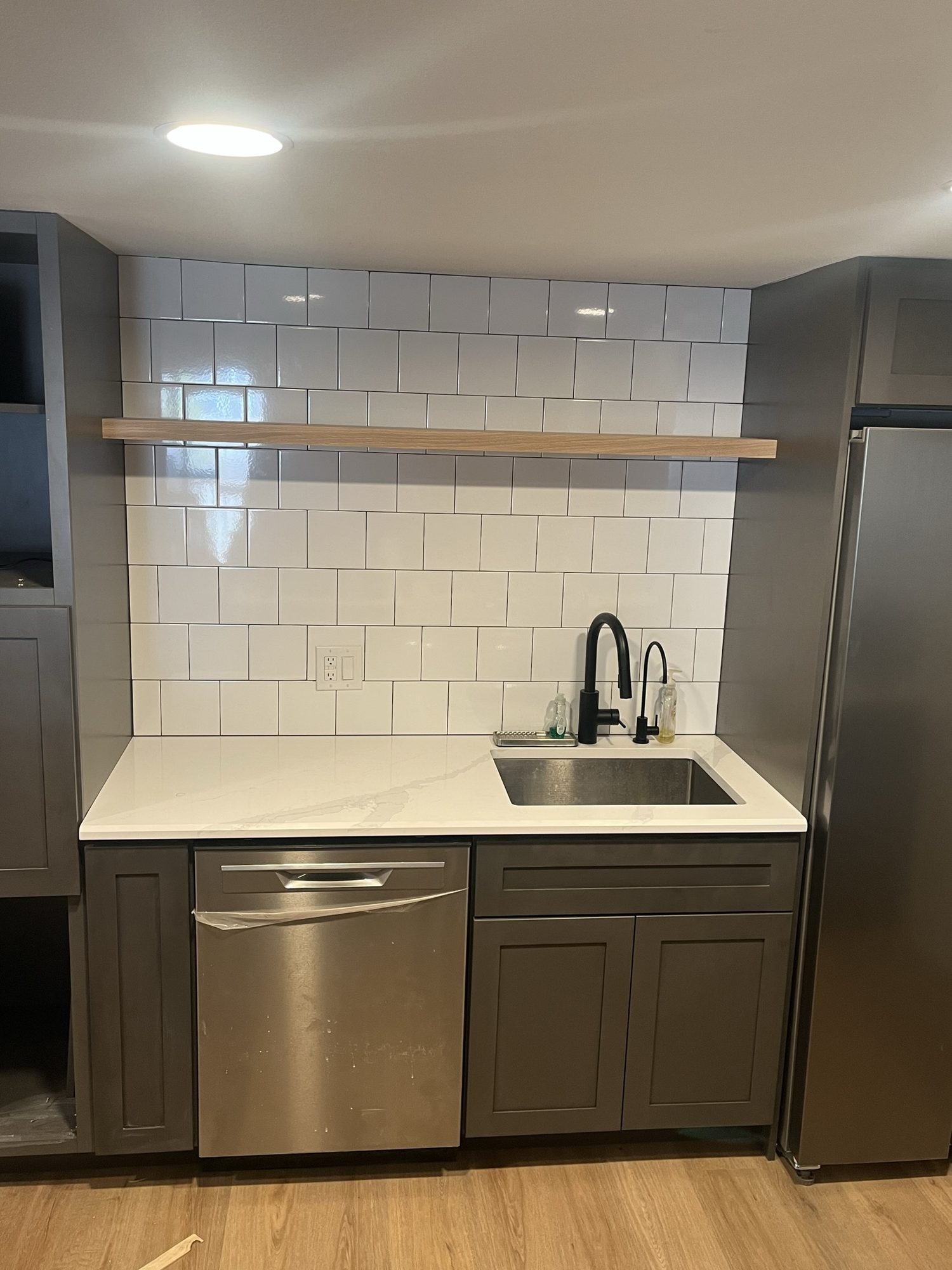 Kitchenette with gray cabinets, white tile backsplash and a wood floating shelf